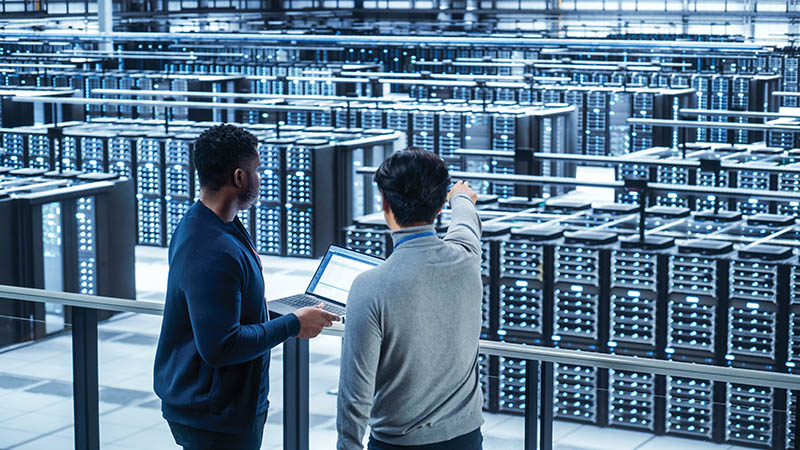 Two Multiethnic Male Data Center System Administrators and IT Specialists Talk, Use Laptop. Information Technology Engineers work on Cyber Security Network Protection in Cloud Computing Server Farm.